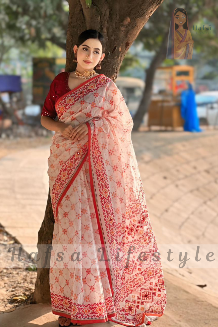 Woman in a white saree with red patterns standing outdoors.